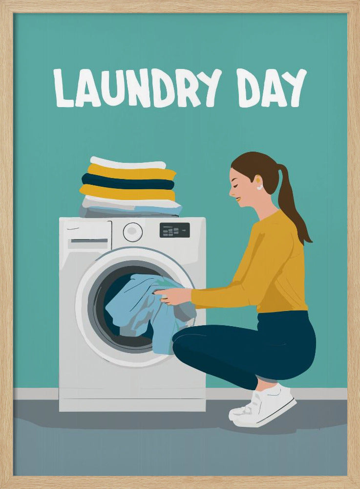 A framed illustration poster with the words 'LAUNDRY DAY' in white at the top. A woman with brown hair in a ponytail, wearing a yellow top and blue pants, crouches to load blue clothes into a white front-loading washing machine. A stack of folded laundry sits atop the machine against a teal background. Print