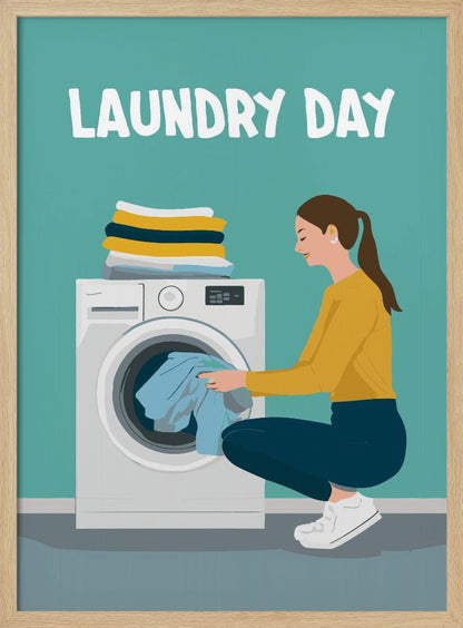 A framed illustration poster with the words 'LAUNDRY DAY' in white at the top. A woman with brown hair in a ponytail, wearing a yellow top and blue pants, crouches to load blue clothes into a white front-loading washing machine. A stack of folded laundry sits atop the machine against a teal background. Print