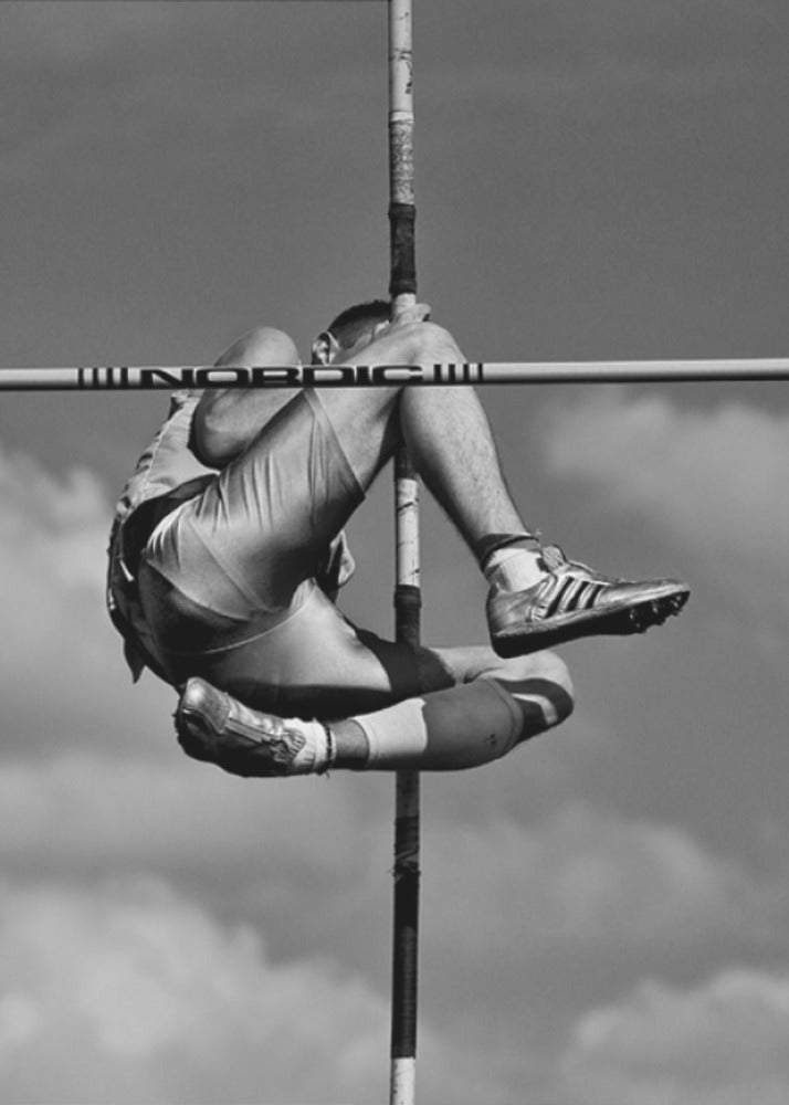 A dramatic black and white, low-angle photograph of a male athlete in mid-air during a pole vault. He is arching his back and bending his legs to clear the horizontal bar, with the vertical pole bisecting the frame. The background is a partly cloudy sky. Wall Art