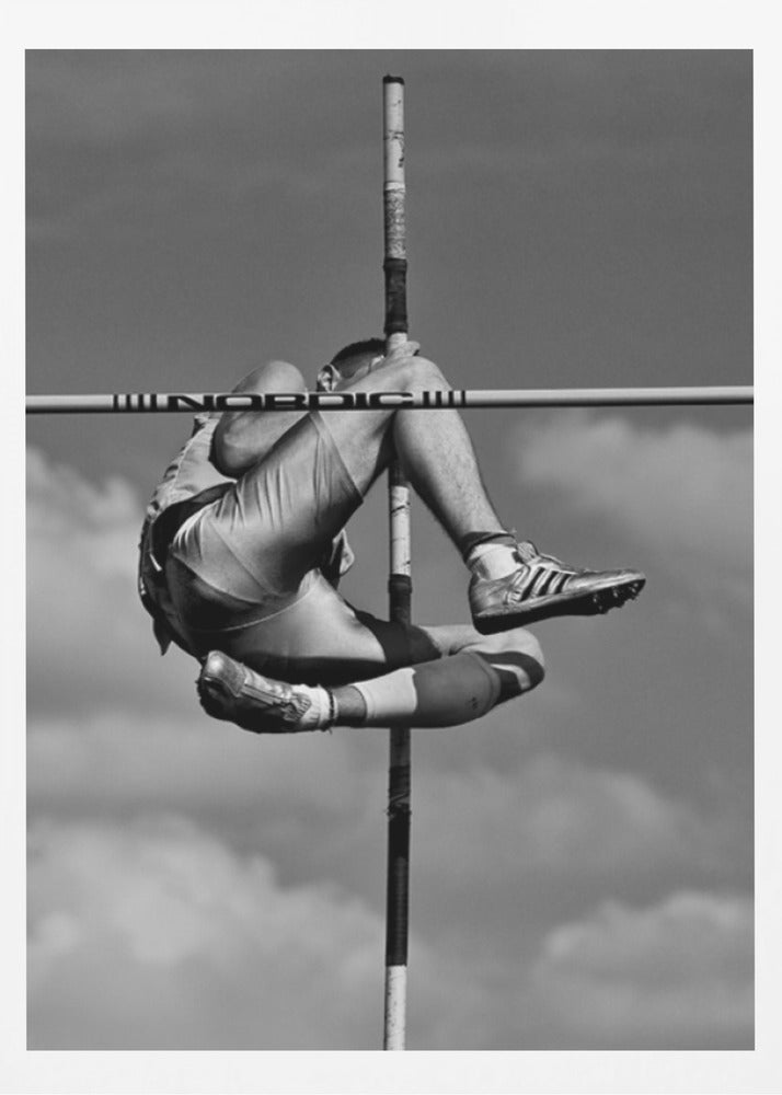 A dramatic black and white, low-angle photograph of a male athlete in mid-air during a pole vault. He is arching his back and bending his legs to clear the horizontal bar, with the vertical pole bisecting the frame. The background is a partly cloudy sky. Wall Art