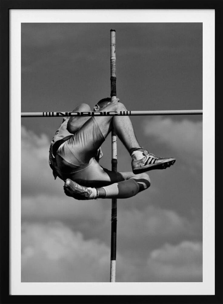 A dramatic black and white, low-angle photograph of a male athlete in mid-air during a pole vault. He is arching his back and bending his legs to clear the horizontal bar, with the vertical pole bisecting the frame. The background is a partly cloudy sky. Wall Art