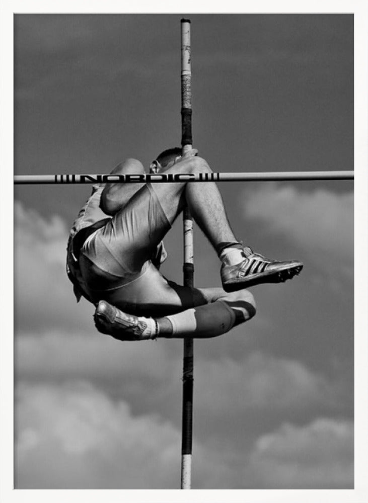 A dramatic black and white, low-angle photograph of a male athlete in mid-air during a pole vault. He is arching his back and bending his legs to clear the horizontal bar, with the vertical pole bisecting the frame. The background is a partly cloudy sky. Wall Art