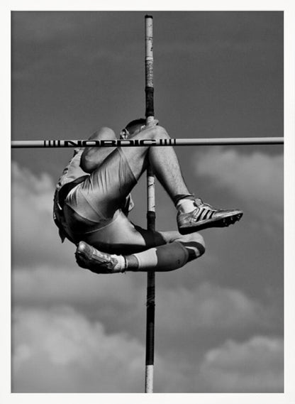 A dramatic black and white, low-angle photograph of a male athlete in mid-air during a pole vault. He is arching his back and bending his legs to clear the horizontal bar, with the vertical pole bisecting the frame. The background is a partly cloudy sky. Wall Art
