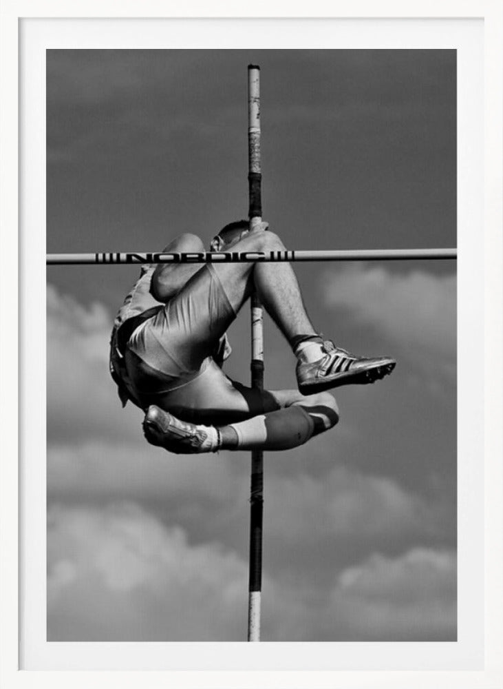 A dramatic black and white, low-angle photograph of a male athlete in mid-air during a pole vault. He is arching his back and bending his legs to clear the horizontal bar, with the vertical pole bisecting the frame. The background is a partly cloudy sky. Wall Art
