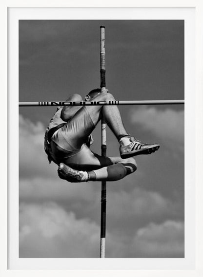 A dramatic black and white, low-angle photograph of a male athlete in mid-air during a pole vault. He is arching his back and bending his legs to clear the horizontal bar, with the vertical pole bisecting the frame. The background is a partly cloudy sky. Wall Art