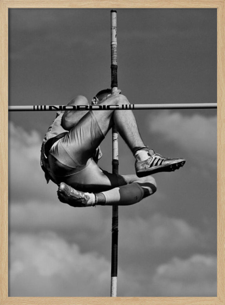 A dramatic black and white, low-angle photograph of a male athlete in mid-air during a pole vault. He is arching his back and bending his legs to clear the horizontal bar, with the vertical pole bisecting the frame. The background is a partly cloudy sky. Wall Art
