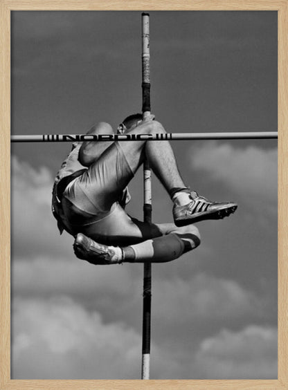 A dramatic black and white, low-angle photograph of a male athlete in mid-air during a pole vault. He is arching his back and bending his legs to clear the horizontal bar, with the vertical pole bisecting the frame. The background is a partly cloudy sky. Wall Art