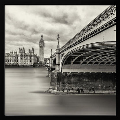 A framed, long-exposure, black and white photograph of London. The image shows Westminster Bridge from a low angle on the right, with the River Thames appearing smooth and misty. In the background on the left are the Houses of Parliament and the Big Ben clock tower under a cloudy sky. Wall Art