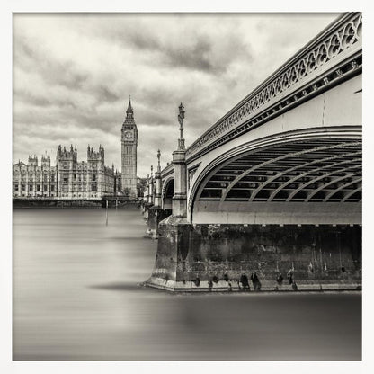A framed, long-exposure, black and white photograph of London. The image shows Westminster Bridge from a low angle on the right, with the River Thames appearing smooth and misty. In the background on the left are the Houses of Parliament and the Big Ben clock tower under a cloudy sky. Wall Art