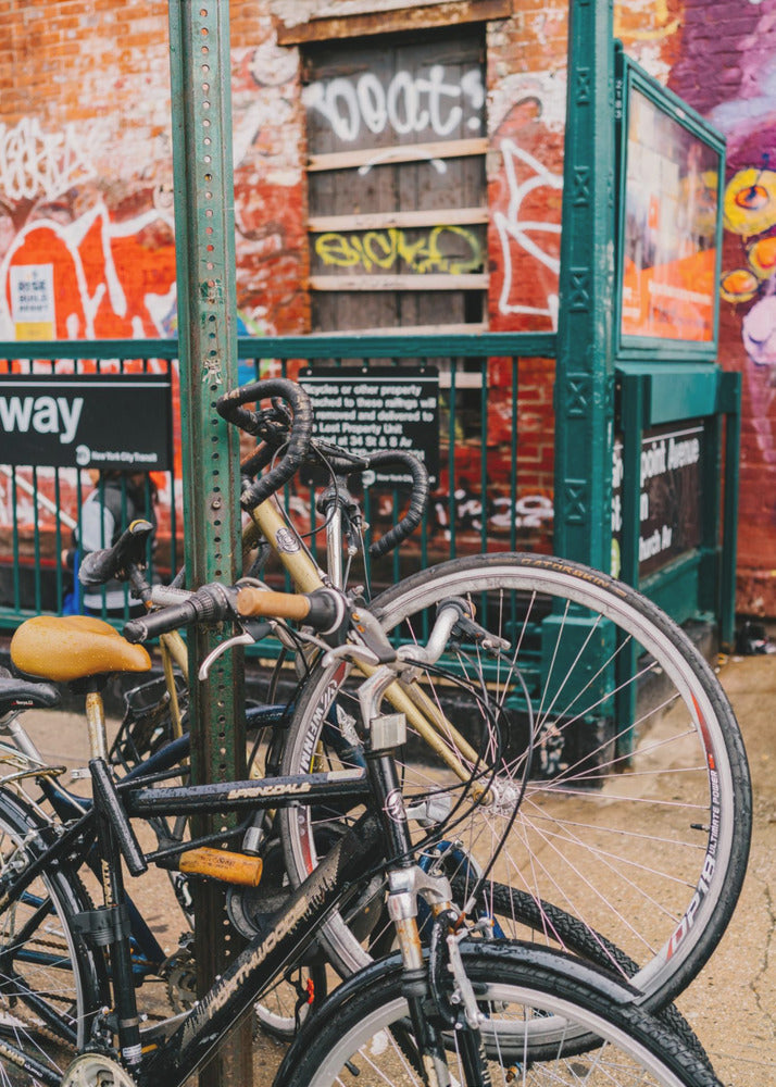 Several bicycles are locked to a green street pole in front of a red brick building covered in graffiti, with a city subway entrance visible in the background. Poster