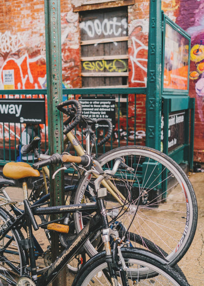 Several bicycles are locked to a green street pole in front of a red brick building covered in graffiti, with a city subway entrance visible in the background. Poster
