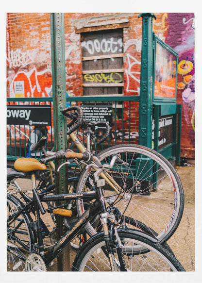 Several bicycles are locked to a green street pole in front of a red brick building covered in graffiti, with a city subway entrance visible in the background. Poster