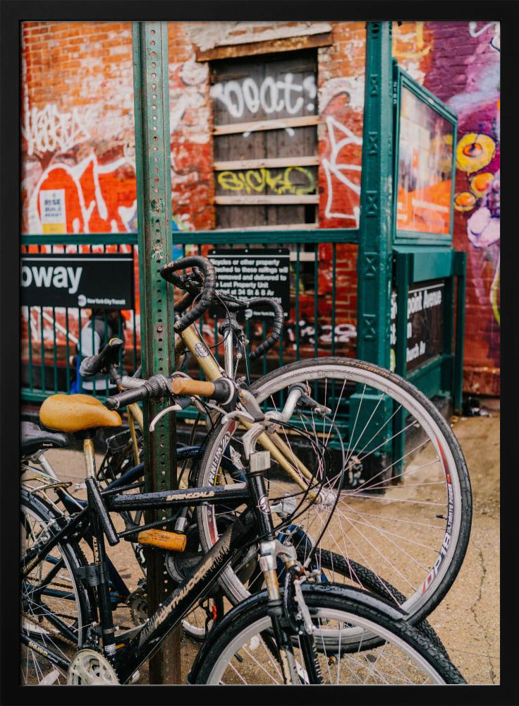 Several bicycles are locked to a green street pole in front of a red brick building covered in graffiti, with a city subway entrance visible in the background. Poster