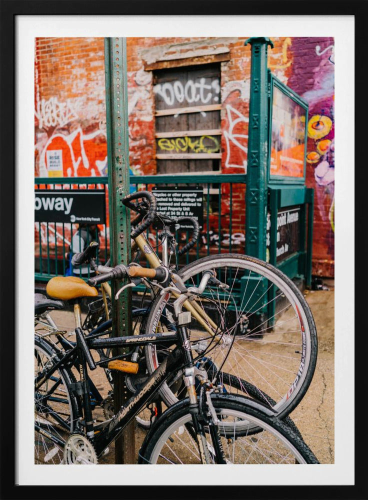 Several bicycles are locked to a green street pole in front of a red brick building covered in graffiti, with a city subway entrance visible in the background. Poster