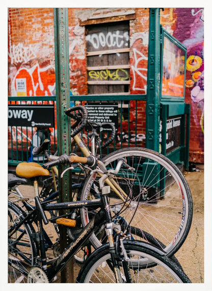 Several bicycles are locked to a green street pole in front of a red brick building covered in graffiti, with a city subway entrance visible in the background. Poster