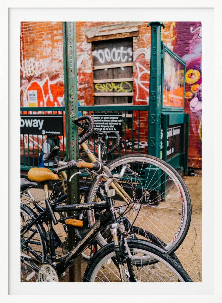 Several bicycles are locked to a green street pole in front of a red brick building covered in graffiti, with a city subway entrance visible in the background. Poster