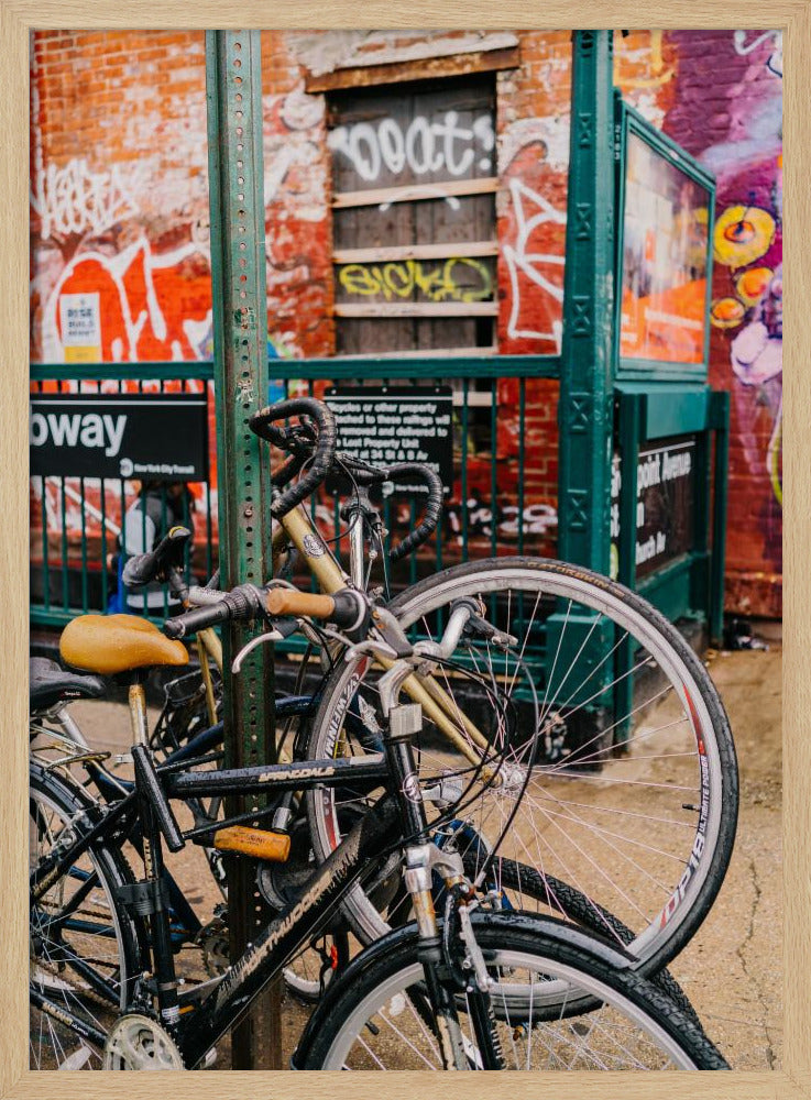 Several bicycles are locked to a green street pole in front of a red brick building covered in graffiti, with a city subway entrance visible in the background. Poster