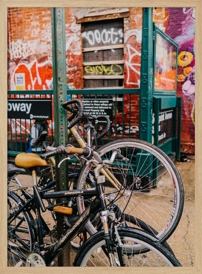 Several bicycles are locked to a green street pole in front of a red brick building covered in graffiti, with a city subway entrance visible in the background. Poster