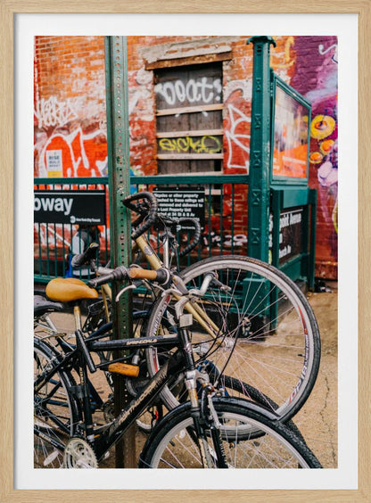 Several bicycles are locked to a green street pole in front of a red brick building covered in graffiti, with a city subway entrance visible in the background. Poster