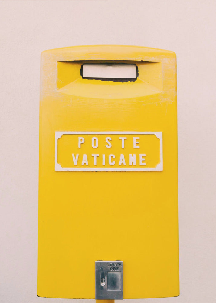 A close-up, front-view photograph of a vibrant yellow Vatican City mailbox against a plain beige wall, enclosed in a thin black frame. The words 'POSTE VATICANE' are printed in white on the front. Artwork