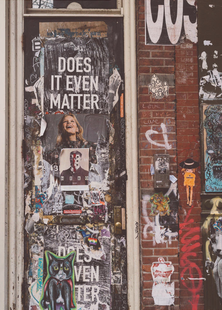 A close-up of a weathered dark door and an adjacent brick wall, both covered in a dense collage of street art, including peeling posters, stickers, and graffiti. A prominent poster on the door asks 'DOES IT EVEN MATTER' in bold white letters. Wall Art