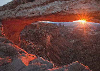 A spectacular sunrise seen through the natural frame of Mesa Arch in Canyonlands National Park, Utah. The sun bursts over the horizon, casting a dramatic orange and red glow on the underside of the rock arch and the vast canyon landscape below. Poster