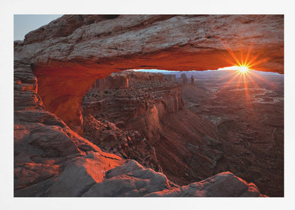A spectacular sunrise seen through the natural frame of Mesa Arch in Canyonlands National Park, Utah. The sun bursts over the horizon, casting a dramatic orange and red glow on the underside of the rock arch and the vast canyon landscape below. Poster