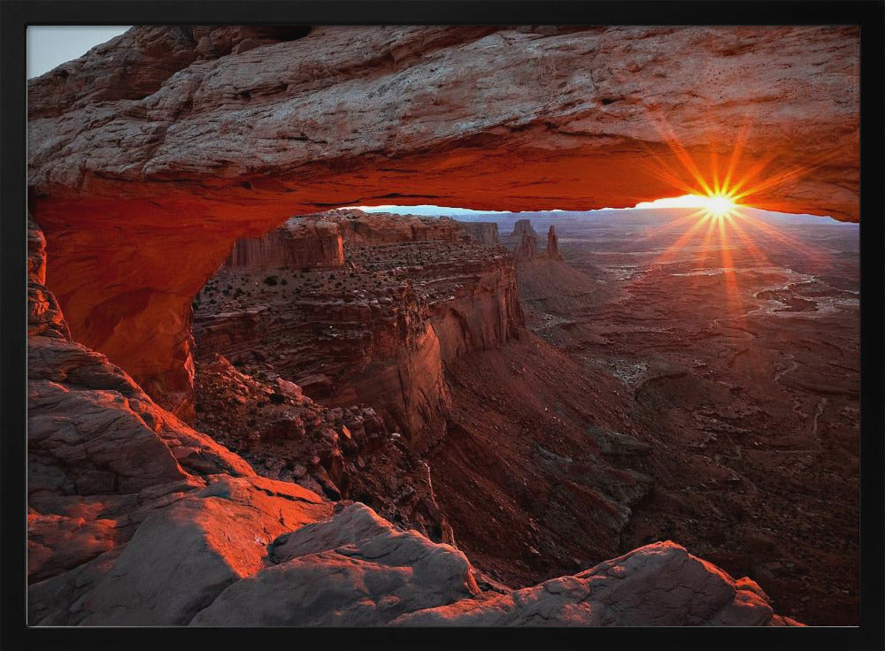 A spectacular sunrise seen through the natural frame of Mesa Arch in Canyonlands National Park, Utah. The sun bursts over the horizon, casting a dramatic orange and red glow on the underside of the rock arch and the vast canyon landscape below. Poster