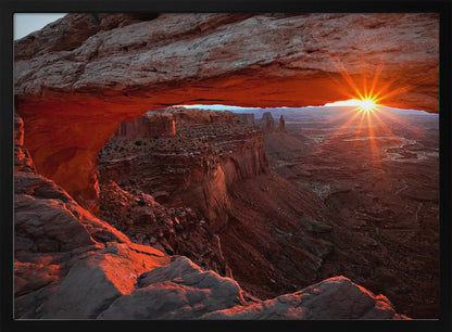 A spectacular sunrise seen through the natural frame of Mesa Arch in Canyonlands National Park, Utah. The sun bursts over the horizon, casting a dramatic orange and red glow on the underside of the rock arch and the vast canyon landscape below. Poster