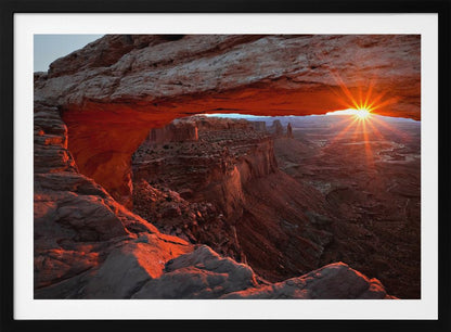 A spectacular sunrise seen through the natural frame of Mesa Arch in Canyonlands National Park, Utah. The sun bursts over the horizon, casting a dramatic orange and red glow on the underside of the rock arch and the vast canyon landscape below. Poster