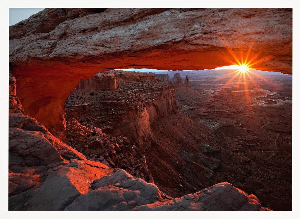 A spectacular sunrise seen through the natural frame of Mesa Arch in Canyonlands National Park, Utah. The sun bursts over the horizon, casting a dramatic orange and red glow on the underside of the rock arch and the vast canyon landscape below. Poster