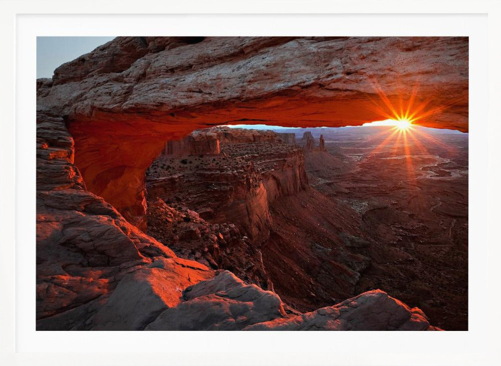 A spectacular sunrise seen through the natural frame of Mesa Arch in Canyonlands National Park, Utah. The sun bursts over the horizon, casting a dramatic orange and red glow on the underside of the rock arch and the vast canyon landscape below. Poster