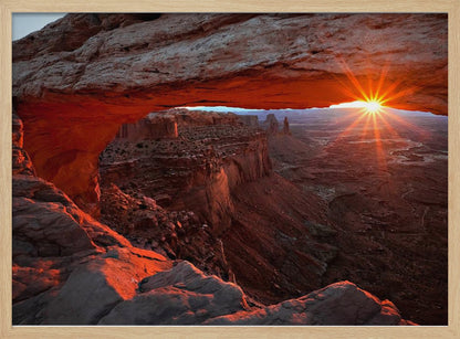 A spectacular sunrise seen through the natural frame of Mesa Arch in Canyonlands National Park, Utah. The sun bursts over the horizon, casting a dramatic orange and red glow on the underside of the rock arch and the vast canyon landscape below. Poster