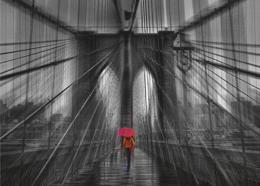 A motion-blurred, black and white photo of the Brooklyn Bridge with a person holding a bright red umbrella walking down the center. The selective color makes the red umbrella the focal point against the dramatic, almost abstract lines of the bridge's cables and arches on a wet, reflective walkway. The image is presented in a silver frame. Wall Art
