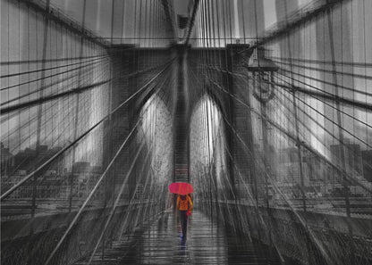 A motion-blurred, black and white photo of the Brooklyn Bridge with a person holding a bright red umbrella walking down the center. The selective color makes the red umbrella the focal point against the dramatic, almost abstract lines of the bridge's cables and arches on a wet, reflective walkway. The image is presented in a silver frame. Wall Art
