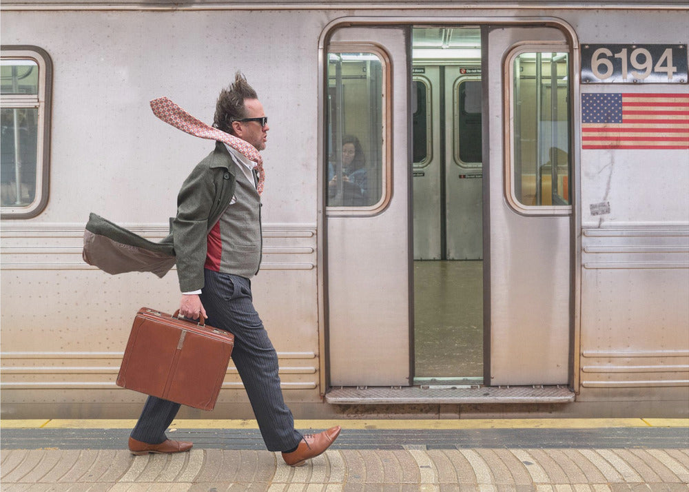 A stylish man with windblown hair and a flying scarf walks briskly along a subway platform, carrying a brown briefcase next to a silver train. Artwork