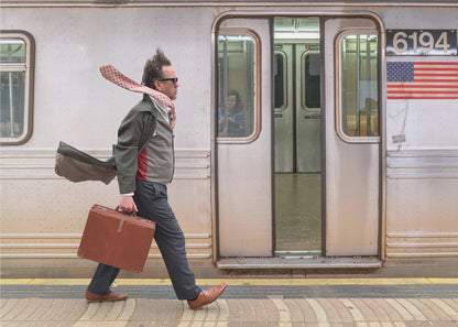 A stylish man with windblown hair and a flying scarf walks briskly along a subway platform, carrying a brown briefcase next to a silver train. Artwork
