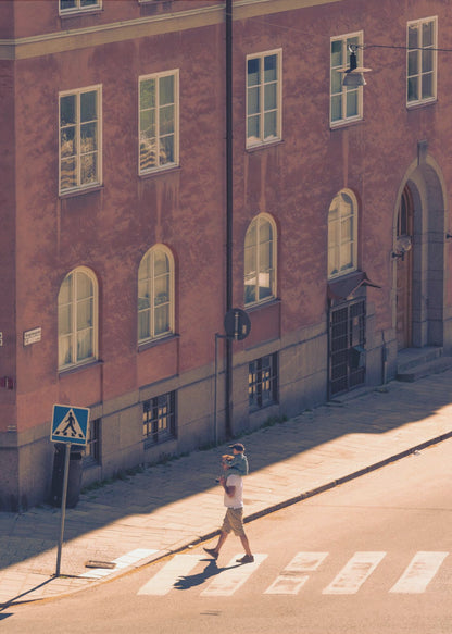 A high-angle photograph of a man carrying a child on his shoulders as they walk across a sunlit crosswalk, with a large, reddish-brown European-style building in the background. The scene is bathed in warm light, creating long shadows. Poster
