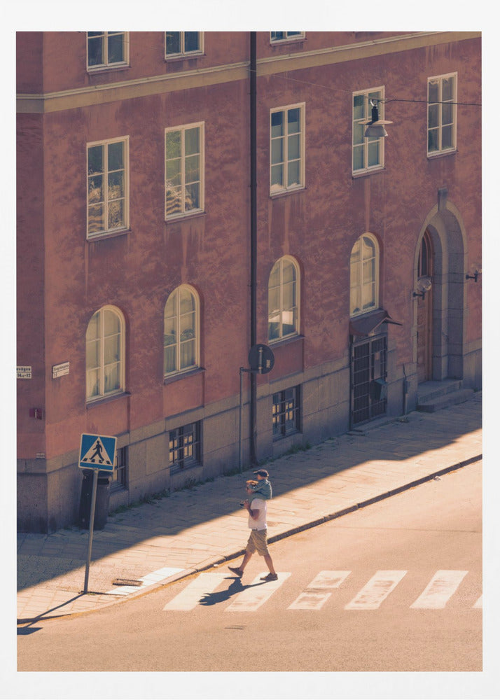 A high-angle photograph of a man carrying a child on his shoulders as they walk across a sunlit crosswalk, with a large, reddish-brown European-style building in the background. The scene is bathed in warm light, creating long shadows. Poster