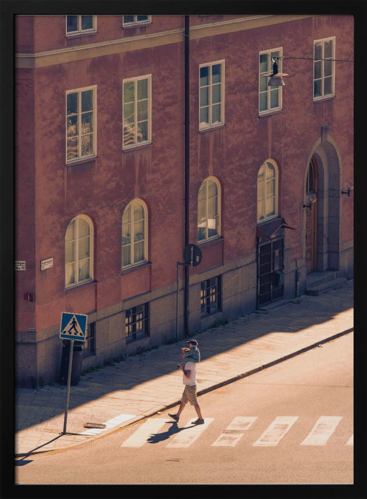 A high-angle photograph of a man carrying a child on his shoulders as they walk across a sunlit crosswalk, with a large, reddish-brown European-style building in the background. The scene is bathed in warm light, creating long shadows. Poster