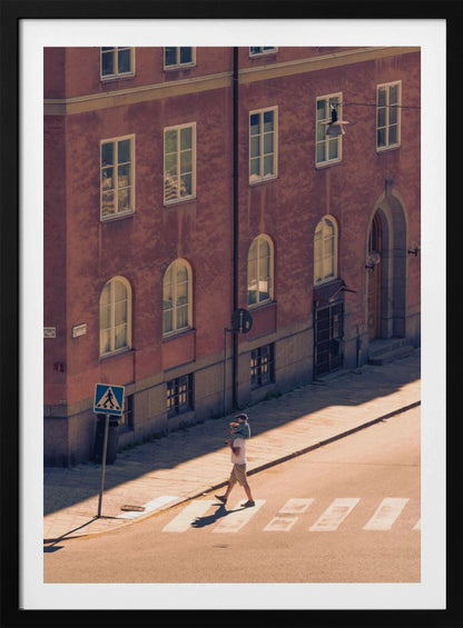 A high-angle photograph of a man carrying a child on his shoulders as they walk across a sunlit crosswalk, with a large, reddish-brown European-style building in the background. The scene is bathed in warm light, creating long shadows. Poster