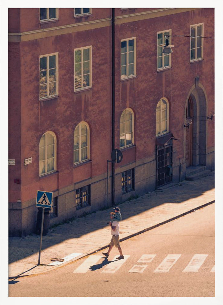 A high-angle photograph of a man carrying a child on his shoulders as they walk across a sunlit crosswalk, with a large, reddish-brown European-style building in the background. The scene is bathed in warm light, creating long shadows. Poster