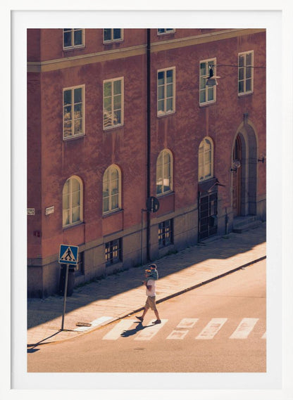 A high-angle photograph of a man carrying a child on his shoulders as they walk across a sunlit crosswalk, with a large, reddish-brown European-style building in the background. The scene is bathed in warm light, creating long shadows. Poster