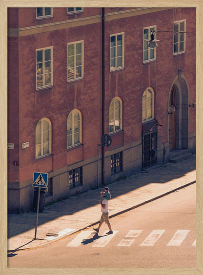 A high-angle photograph of a man carrying a child on his shoulders as they walk across a sunlit crosswalk, with a large, reddish-brown European-style building in the background. The scene is bathed in warm light, creating long shadows. Poster