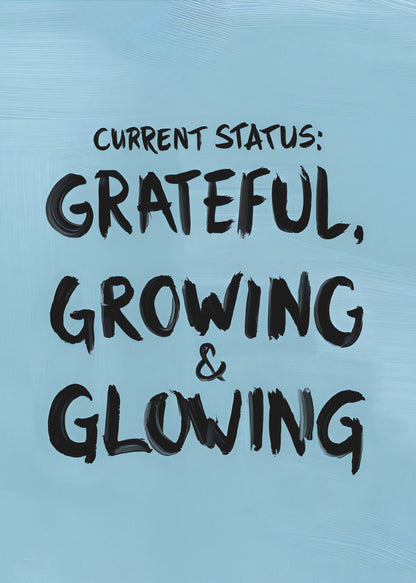A motivational poster with a light blue background and a thin black frame. The text 'CURRENT STATUS: GRATEFUL, GROWING & GLOWING' is written in a bold, black, handwritten brush font. Decor