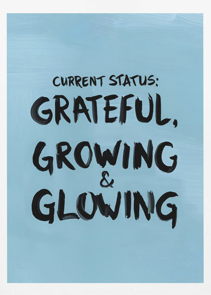 A motivational poster with a light blue background and a thin black frame. The text 'CURRENT STATUS: GRATEFUL, GROWING & GLOWING' is written in a bold, black, handwritten brush font. Decor