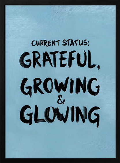 A motivational poster with a light blue background and a thin black frame. The text 'CURRENT STATUS: GRATEFUL, GROWING & GLOWING' is written in a bold, black, handwritten brush font. Decor
