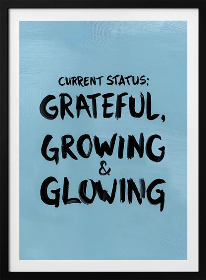 A motivational poster with a light blue background and a thin black frame. The text 'CURRENT STATUS: GRATEFUL, GROWING & GLOWING' is written in a bold, black, handwritten brush font. Decor