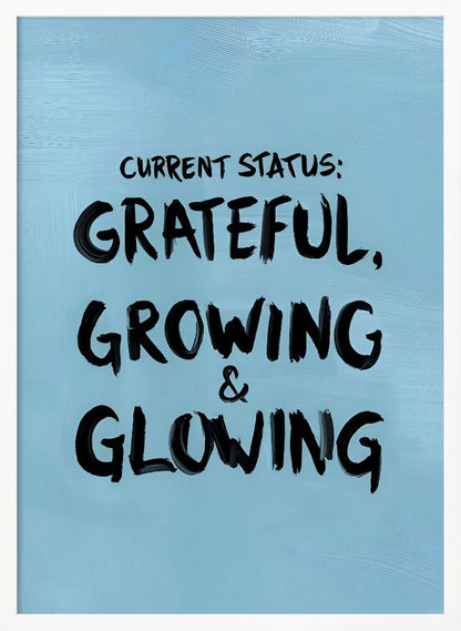 A motivational poster with a light blue background and a thin black frame. The text 'CURRENT STATUS: GRATEFUL, GROWING & GLOWING' is written in a bold, black, handwritten brush font. Decor