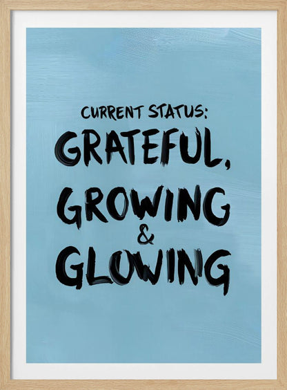 A motivational poster with a light blue background and a thin black frame. The text 'CURRENT STATUS: GRATEFUL, GROWING & GLOWING' is written in a bold, black, handwritten brush font. Decor